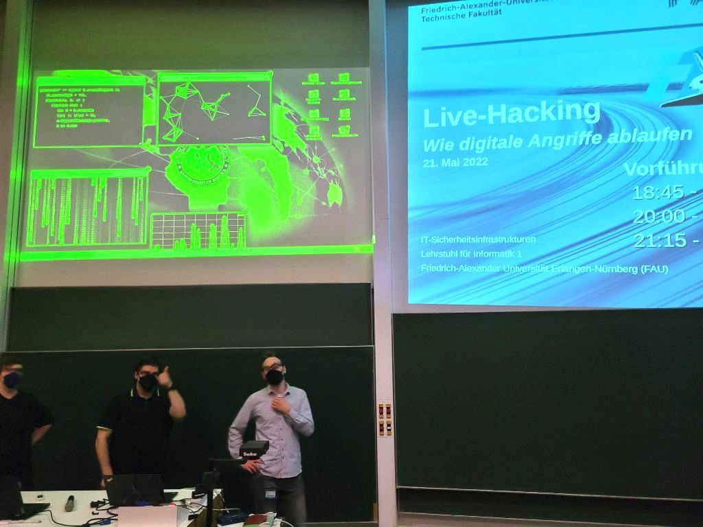 Davide and 2 colleagues wearing black masks in front of event room. In the background there are presentation slides of the live hacking event.